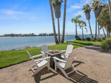 A lakeside patio with a lit fire pit and white Adirondack chairs under tall palm trees by the water.
