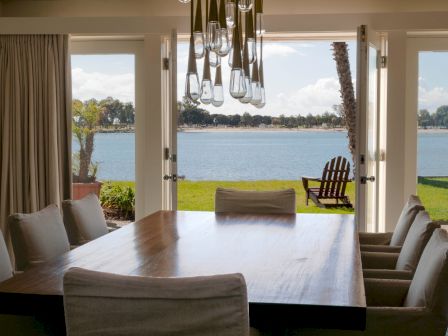 A bright dining room with a large table and cushioned chairs, looking out through open doors onto a calm lake and lawn.