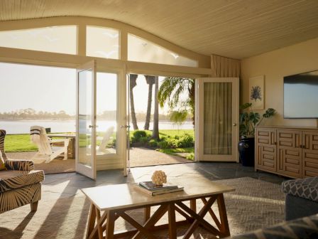 A bright living room with open French doors to a sunny patio and lawn by the water, with couches, coffee table, and wall TV.