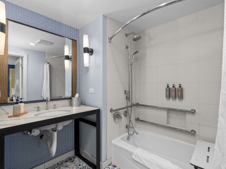 Modern bathroom with blue-tiled vanity, large mirror, bathtub with hand shower, towel, and wall toiletries.