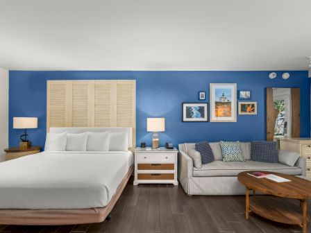 A bright bedroom with a white bed, blue accent wall, framed ocean art, lamps, and a gray couch with patterned pillows.
