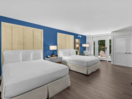 A bright hotel bedroom with two white beds, blue accent wall, shutters, lamps, and patio doors to greenery outside.