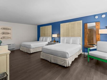 A bright blue hotel room with two white beds, shutter windows, a mirror, and a green accent chair.