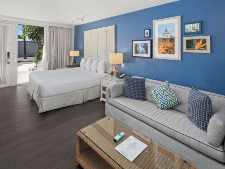 A bright hotel-style bedroom with a white bed, gray sofa, blue walls, lamps, and framed ocean photos.