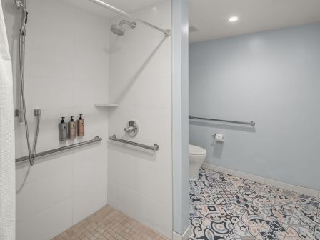 A modern bathroom showing a walk-in shower with fixtures, bottles, patterned tile floor, and a toilet with towel rail.