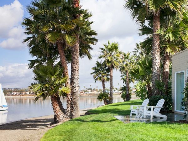 A lakeside patio with green lawn, white lounge chairs, tall palm trees, and a sailboat on calm water under a blue sky.