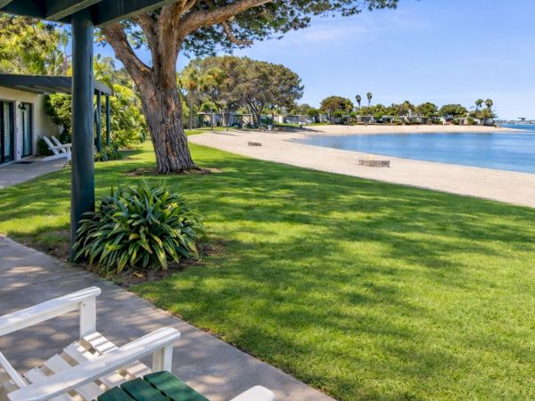 A sunny beachfront resort: green lawn, shaded walkway, white seating, and a calm sandy shore beside blue water.