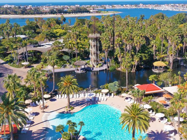 Aerial view of a tropical resort with a sparkling pool, palm trees, a lake, and the ocean beyond under sunny skies.
