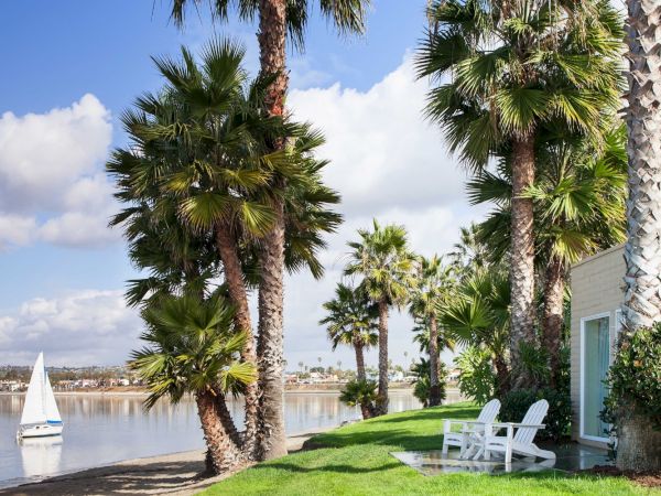 Lakeside garden with tall palm trees, a green lawn, white chairs, and a sailboat on calm water under blue skies.