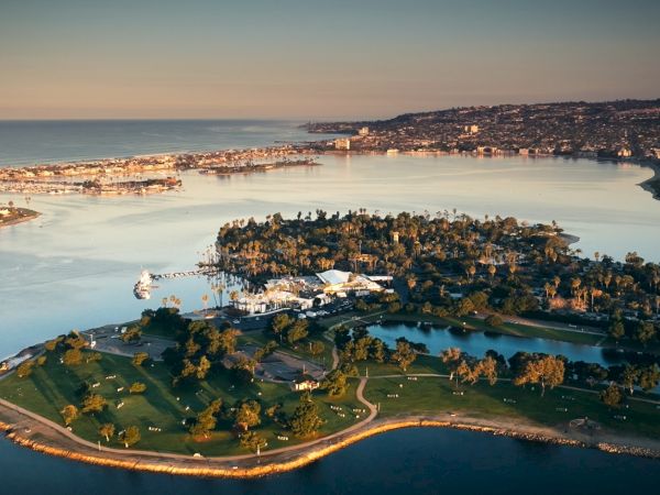 Aerial view of a coastal bay at sunset with a park island, harbor lights, and hillside homes along the shoreline.