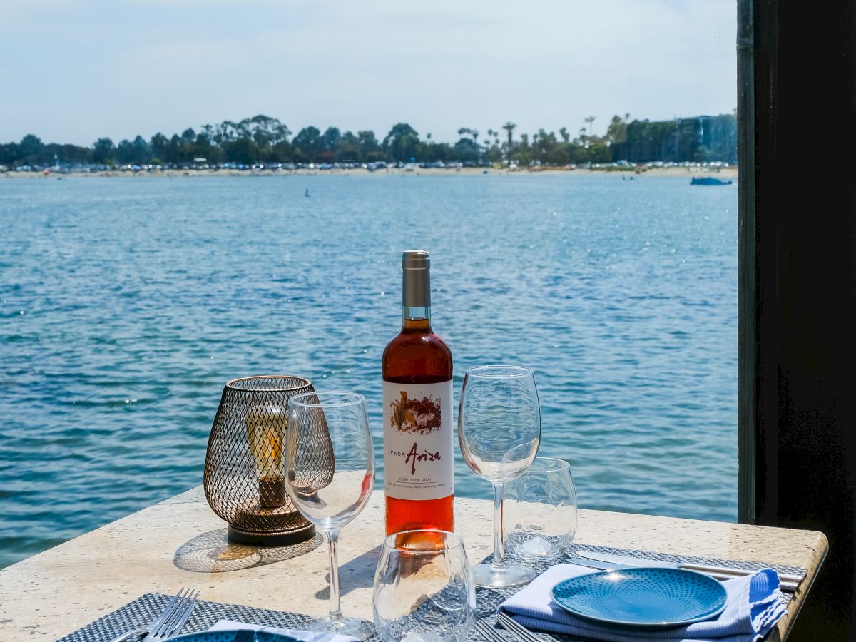 A waterfront table set with a wine bottle, glasses, and plates, overlooking a calm river under a blue sky.