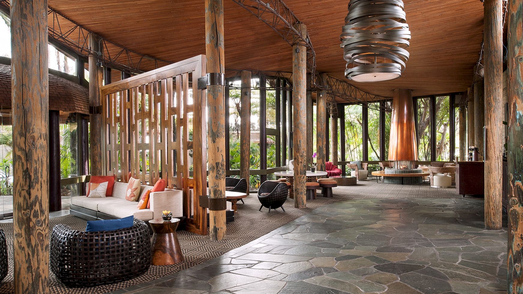 A rustic outdoor lounge with wooden posts, woven seating, patterned floor tiles, and a hanging chandelier-style light under a canopy.