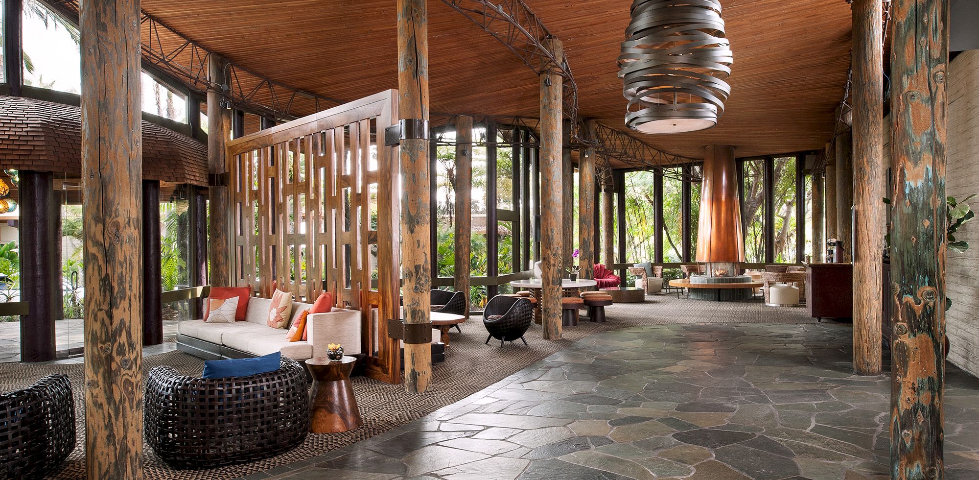 A rustic outdoor lounge with wooden posts, woven seating, patterned floor tiles, and a hanging chandelier-style light under a canopy.