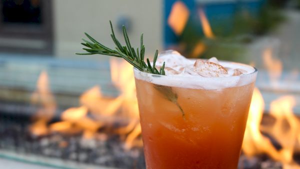 A tall glass of iced orange drink garnished with a sprig of rosemary, outdoors by a pool.