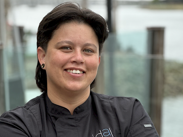 A smiling chef in a black uniform with &ldquo;Charlotte Guerrero Chef De Cuisine&rdquo; on the chest, arms crossed.