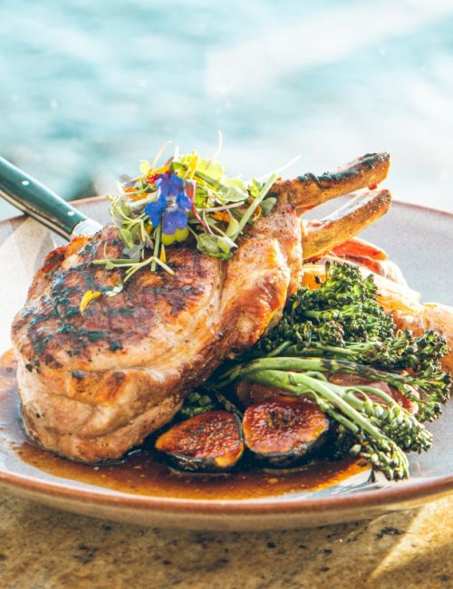 A seaside plate of grilled lamb chops topped with microgreens, broccoli, and sauce beside the ocean.