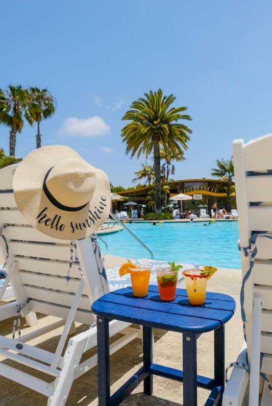 A blurry poolside scene with palm trees, stone walls, and a set of colorful items by a blue swimming pool.