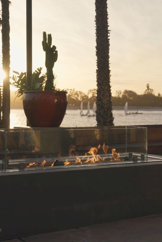 A beachside patio at sunset with a lit outdoor fire pit, palm trees, and water in the background.