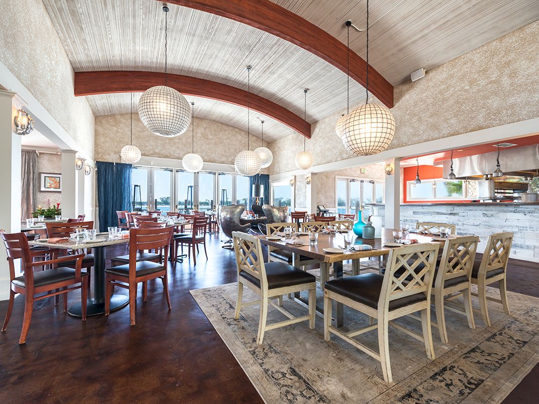 A bright restaurant dining area with long tables, wooden chairs, and large globe pendant lights above an open kitchen buffet.