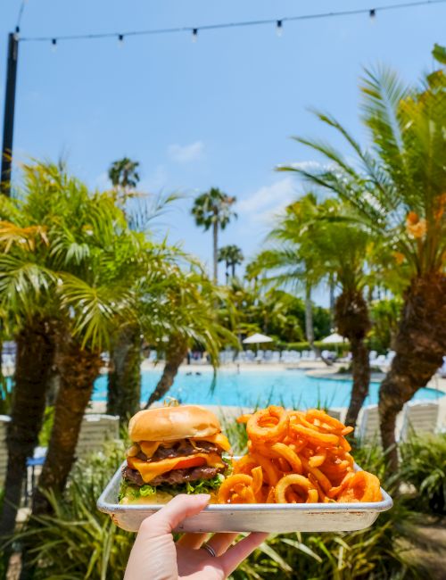 Poolside palms with a hand holding a tray: a burger and a pile of orange fries, against a bright blue sky, nearby.