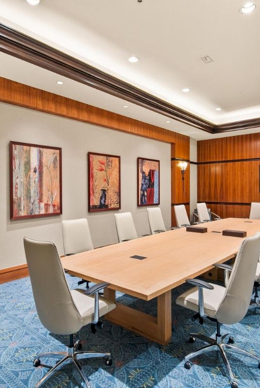 A modern conference room with a long light-wood table, swivel chairs, framed art, and warm wooden walls under ceiling lights.