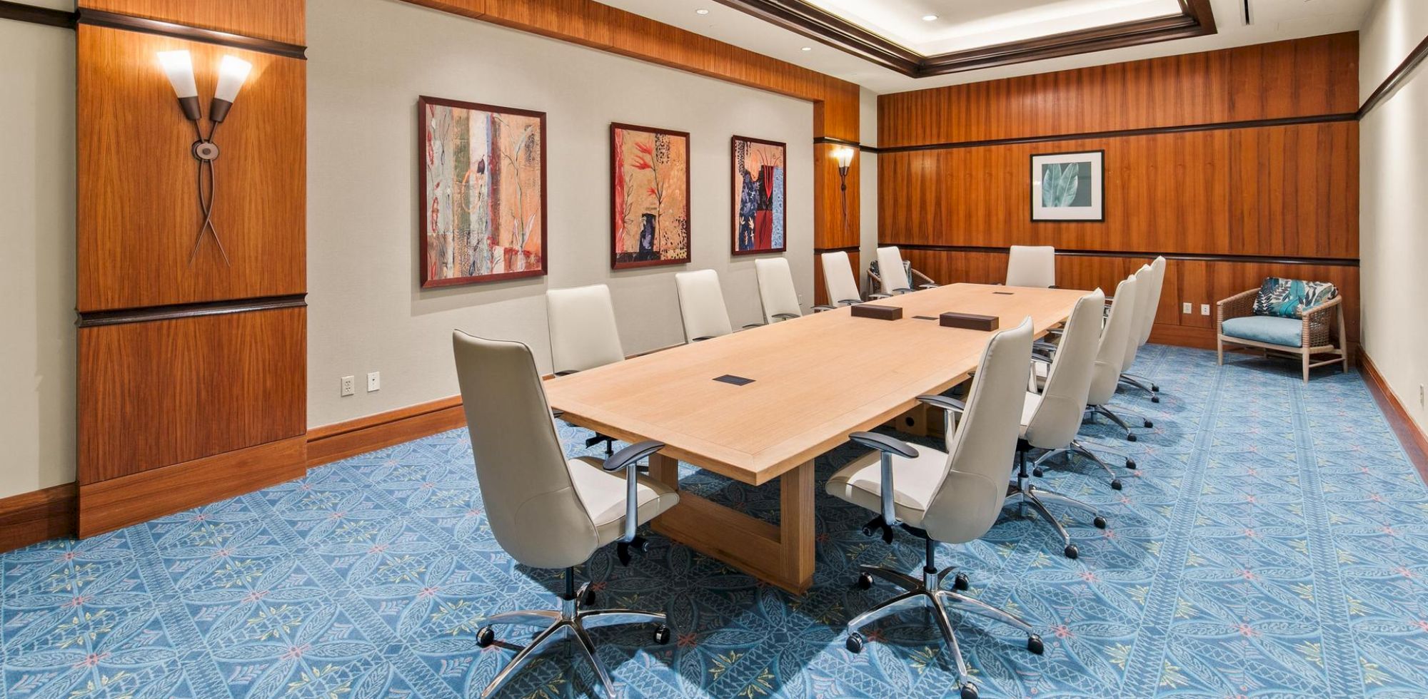 A modern conference room with a large wood table, white chairs, framed art, and blue patterned carpet.
