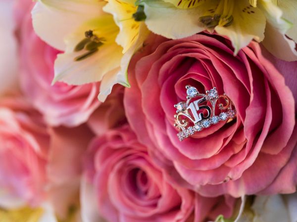Close-up of pink roses with white/yellow blossoms, butterflies, and a sparkly silver jeweled letter charm nestled in the petals.