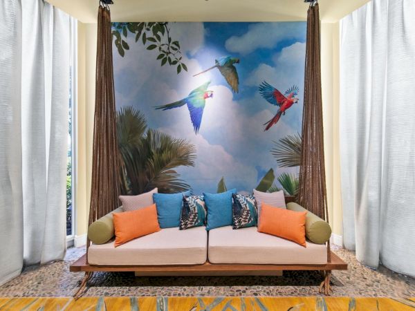 A tropical lounge: two-seater sofa with bright pillows beneath a colorful mural of flying parrots and palm trees.