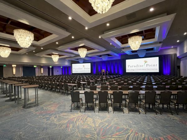 An empty conference room at Paradise Point with rows of chairs, chandeliers, and a stage lit in blue and purple, ready for events.