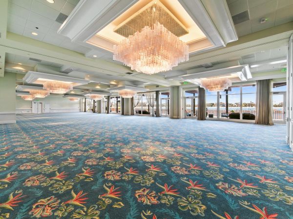 A large event hall with blue patterned carpet and sparkling crystal chandeliers, with doors open to a bright waterfront view.