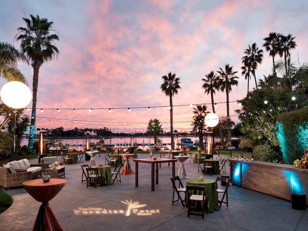 An outdoor event setup at sunset with palm trees, string lights, and tables/chairs by a marina and poolside.