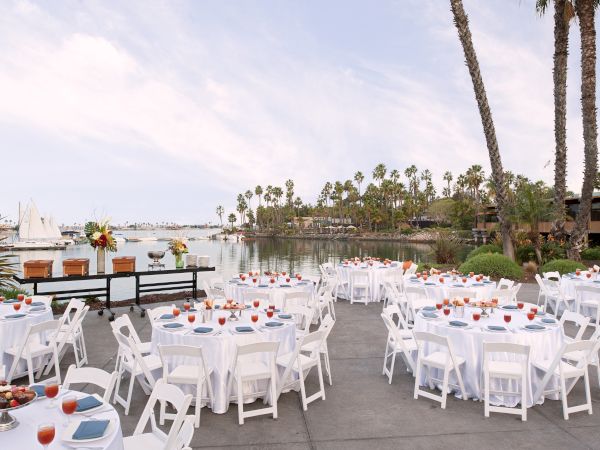 Outdoor waterfront banquet: white tables set with place settings, orange drinks, palm trees, and sailboats under a cloudy sky.