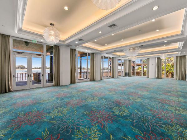An elegant hotel conference room with blue patterned carpet, chandeliers, tall windows overlooking water, and an EXIT sign.