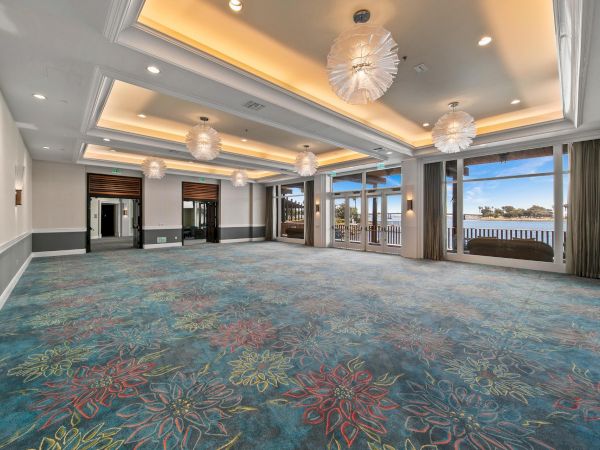 An elegant hotel event room with blue patterned carpet, tall windows overlooking the water, and ornate hanging chandeliers.