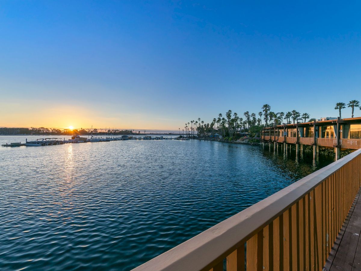 Sunset over a calm marina, with boats, palm-lined shore, and a wooden pier stretching into the golden sky.