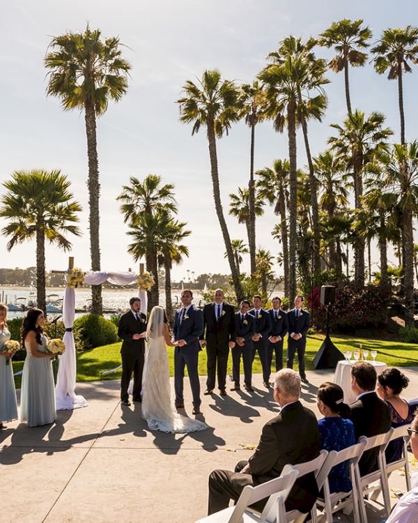 Outdoor seaside wedding with palm trees, bridesmaids in white and groomsmen in suits standing before the couple.