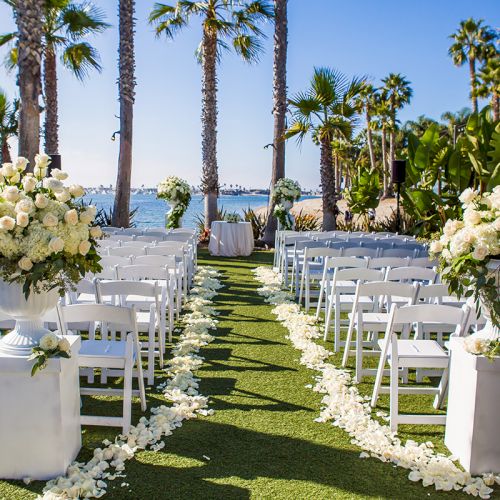 An outdoor wedding aisle lined with white chairs, lush white flowers in vases, and tall palm trees by the ocean.