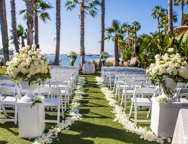 An outdoor wedding aisle lined with white chairs, lush white flowers in vases, and tall palm trees by the ocean.