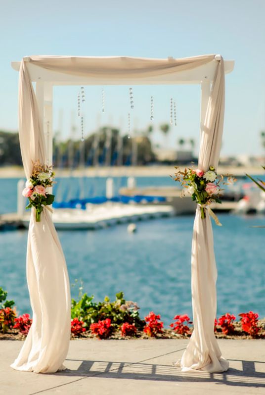 A seaside wedding ceremony setup: a white floral arch over a walkway, with chairs, bouquets, and boats in the background.