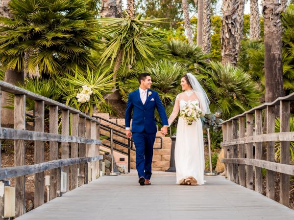 A bride and groom in wedding attire stroll hand-in-hand on a wooden bridge surrounded by palm trees and greenery.