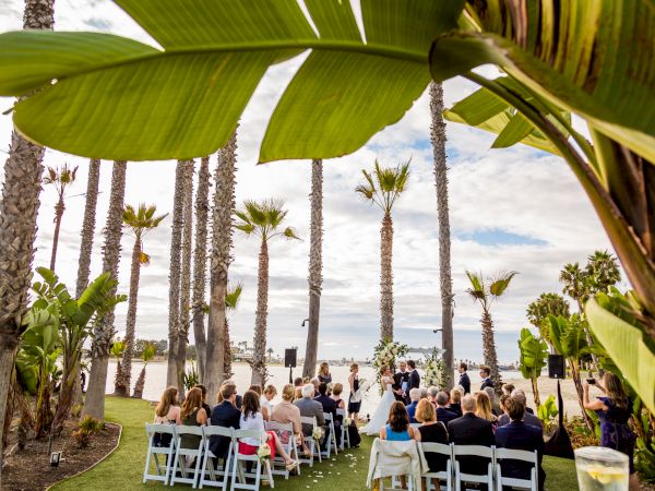 An outdoor wedding ceremony under lush green palm-leaf canopies, with guests seated on chairs near the waterfront.