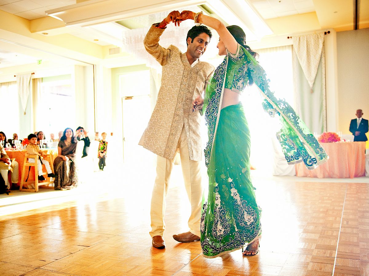 A couple dances at a celebration, the man in a traditional beige outfit and the woman in a green embroidered dress, smiling joyfully.
