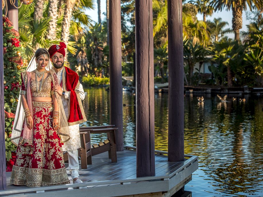 A dressed-up wedding couple stands on a pier by a calm lake, framed by tall wooden pillars and lush palm trees.