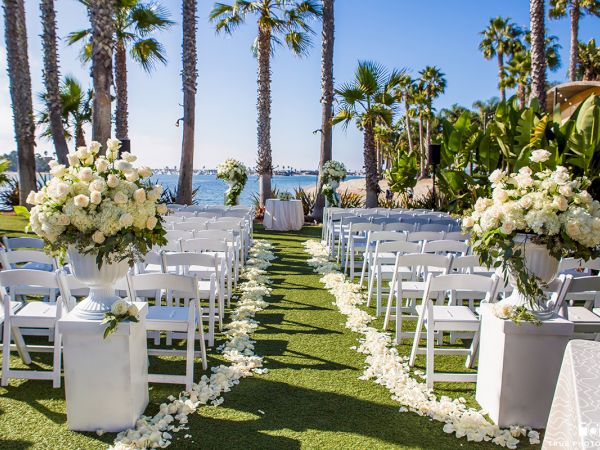 Beach wedding aisle with white chairs, bouquets and floral decor under tall palm trees by the ocean.