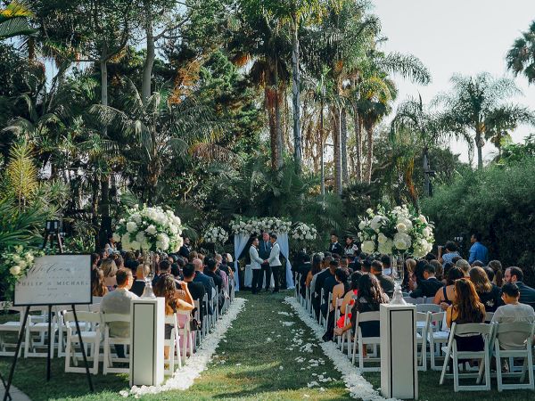 Outdoor wedding ceremony in a lush garden, with a flower-lined aisle, chairs filled with guests, and a couple standing at the front.