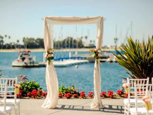 A wedding ceremony setup with a floral arch on a waterfront dock, white chairs, and boats in the background.