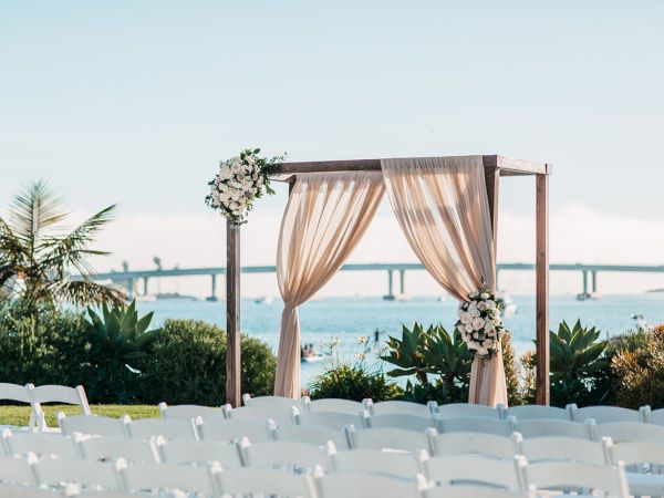 An outdoor wedding ceremony setup: white chairs, floral arch, and flowing fabric beside the sea with a bridge in view.