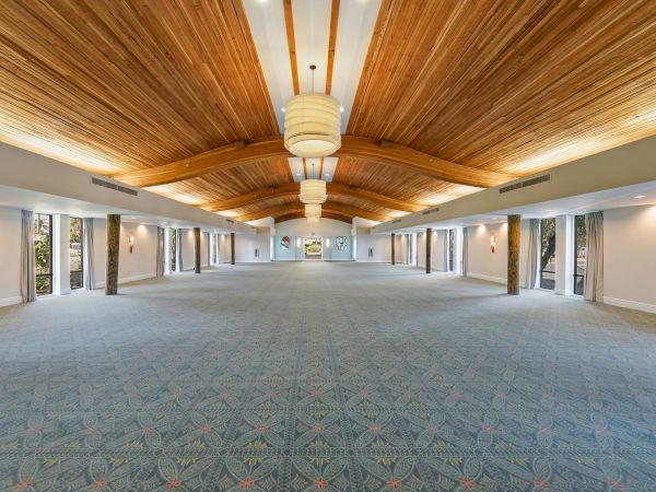 An elegant indoor hall with a patterned carpet, tall windows, and a vaulted wood ceiling with hanging light fixtures and pillars.