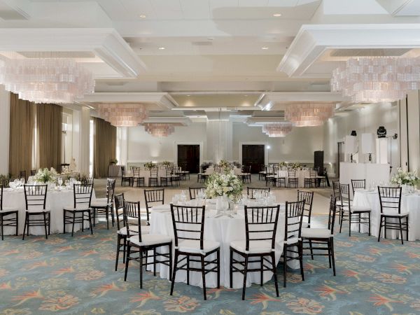 A decorated banquet hall with tables, black chairs, white linens, and floral centerpieces under hanging chandeliers.