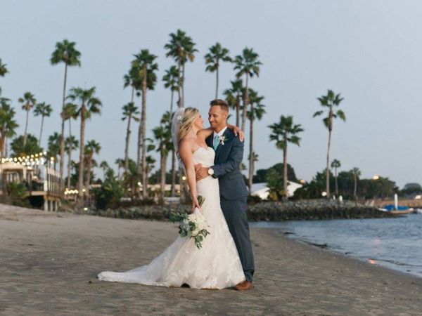 A bride and groom in wedding attire pose on a palm-lined beach at dusk, smiling and holding each other.